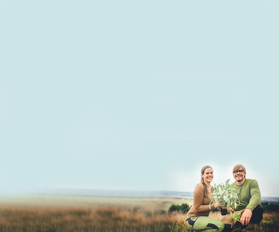 Marie und Nils Schneider hocken nebeneinander vor weitläufiger Landschaft und halten Gartenpflanzen in den Händen.