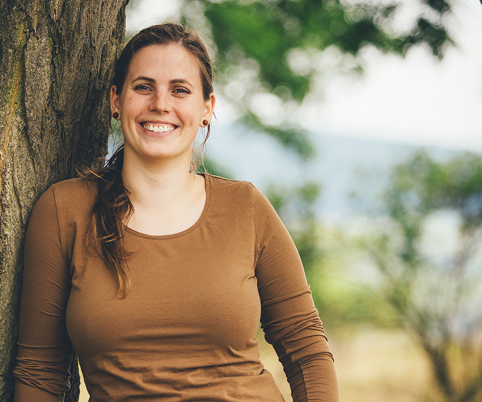 Portrait von Marie Schneider. Sie steht lächelnd angelehnt an einen Baum.